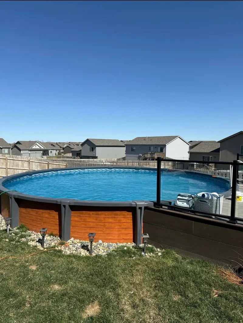 Round above-ground pool with a wood-look wall, a deck with glass railing and landscape lights in a newer neighborhood